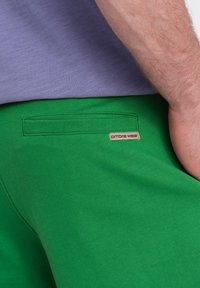 Un pantalon de survêtement vert en coton doux, avec une petite étiquette beige portant l'inscription "ombre wear", et une poche arrière avec une finition côtelée.