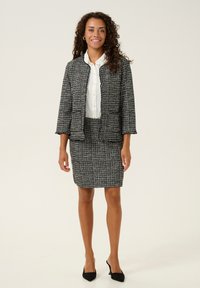 Fekete-fehér tweed zakó és szoknya szett, amely texturált anyagból készült, kockás mintával. Fehér blúz fölött viselve, fekete magassarkúval párosítva.