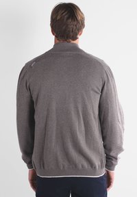 Homme aux cheveux bruns courts portant un pull gris à manches longues et un pantalon sombre, se tenant de dos contre un arrière-plan clair et uni.