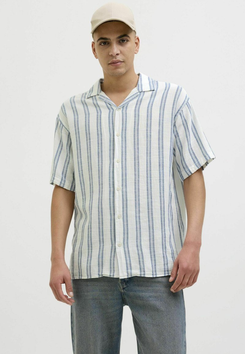 Chemise boutonnée en tissu léger avec des rayures verticales bleues et blanches, à manches courtes, et un col cranté. Associée à un jean en denim clair.
