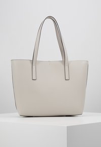 Beige tote bag med en glat overflade, med to flade håndtag og kontrasterende sort kant langs toppen.