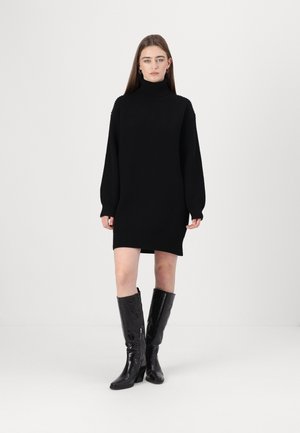 Schwarzes gestricktes Kleid mit hohem Rollkragen, fallenden Schultern und lockerer Passform, kombiniert mit knielangen, glänzend schwarzen, hohen Stiefeln.