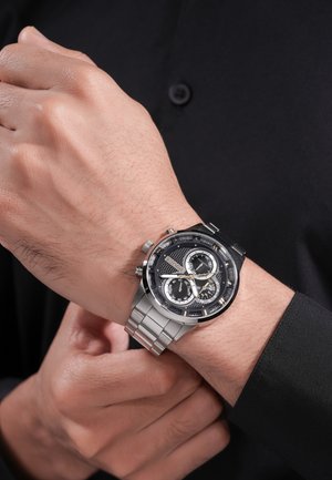 Person, der justerer et sølvfarvet metalarmbåndsur med sort urskive med flere skiver, iført en sort langærmet skjorte.