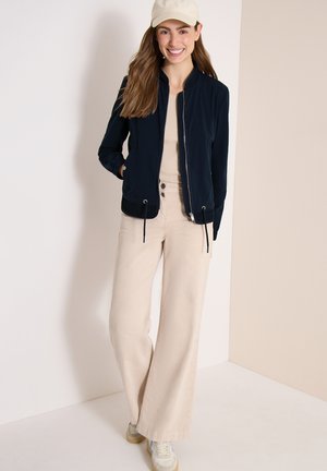 Jeune femme portant une casquette beige, une veste à fermeture éclair noire, un pantalon beige à jambes larges et des baskets, souriant et posant devant un mur bicolore beige.