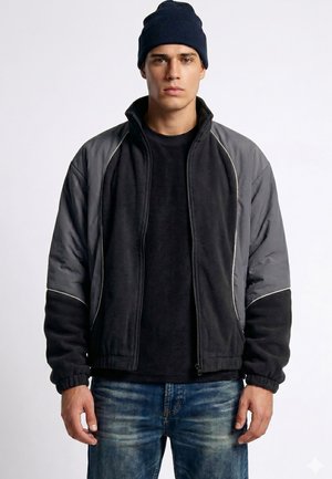 Jeune homme portant un bonnet noir, une veste zippée noire et grise, un t-shirt noir et un jean bleu, debout devant un fond blanc uni.