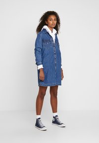 Denimist shirtkleit nupudega ees, kahe rinna taskuga, pikkade varrukatega ja voodrita viimistlusega. Kantud valge kapuutsiga särgi kohal ja koos tumesiniste tennisekingadega.