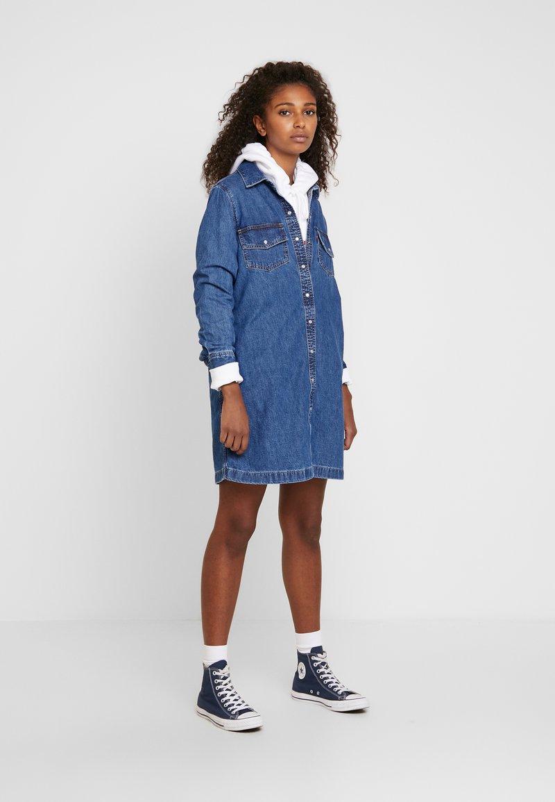 Robe chemise en denim avec un devant à boutons, deux poches poitrine, des manches longues et une finition non doublée. Portée sur un sweat à capuche blanc et associée à des baskets bleu marine.