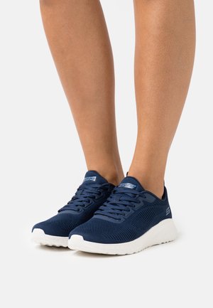 Jambes portant des baskets de sport en mesh bleu marine avec des semelles blanches, debout sur un fond clair uni.