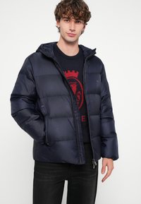 Marineblaue Steppjacke mit Kapuze, ausgestattet mit Reißverschluss vorne, gesteppter Optik und glatter Textur. Getragen über einem dunklen Pullover mit rotem Emblem.