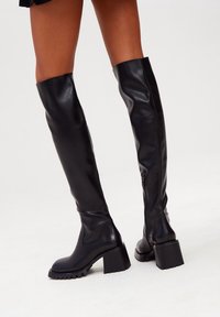 Botas de cuero negras por encima de la rodilla con tacón ancho, que cuentan con una cremallera lateral y una textura suave. Suela de goma para mayor agarre.