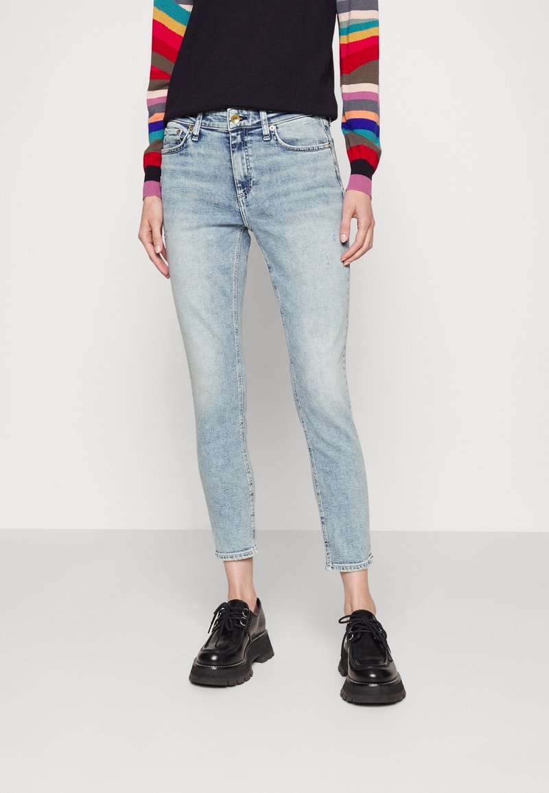 Jean bleu clair ajusté avec une légère élasticité, doté d'un design classique à cinq poches et d'un ourlet effiloché, accompagné de chaussures noires à lacets.
