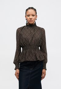 Brun mønstret bluse med høy hals, smokked midje og lange vide ermer, kombinert med et mørkt denimpanel.