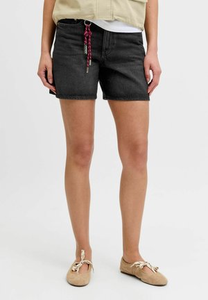 Person iført sorte denimshorts med rød reb-nøglering, beige jakke og beige balletsko med mønstrede bånd.