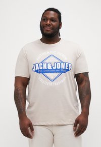 Gaiši bež T-krekls ar zilu un baltu grafisku printu ar "Jack & Jones" logotipu, īsām piedurknēm, apaļu kakla izgriezumu un mīkstu tekstūru.