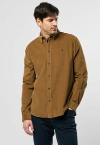 Brun kordfløjlsskjorte med krave og knaplukning foran, samt et lille broderet logo på brystet, parret med mørke jeans.