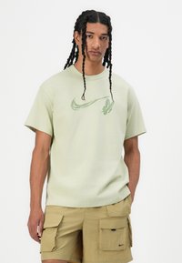 Light green cotton t-shirt with a small green Nike logo. Worn with tan cargo shorts featuring multiple pockets and black accents.