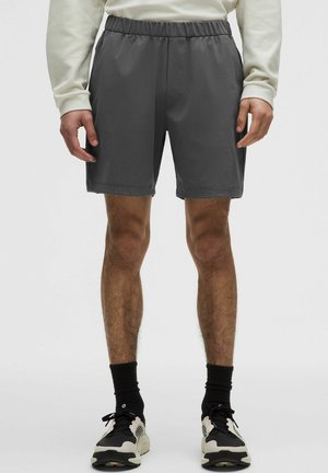 Mand iført mørkegrå træningsshorts, cremefarvet langærmet skjorte, sorte sokker og sort-hvide sneakers, stående mod en ensfarvet baggrund.