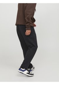 Tmavé tepláky s volným střihem a bočními kapsami. Nošeny se hnědou mikinou a černo-bílými teniskami s modrými akcenty.