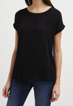 Femme portant un chemisier noir uni à manches courtes avec manches retroussées et un jean bleu sur un fond uni.