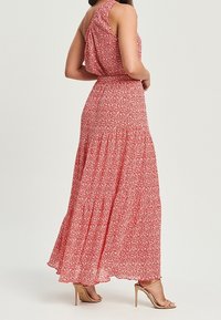 Robe maxi en tissu rouge avec un motif floral, sans manches, taille cintrée et jupe à volants. Associée à des chaussures à talons nudes.