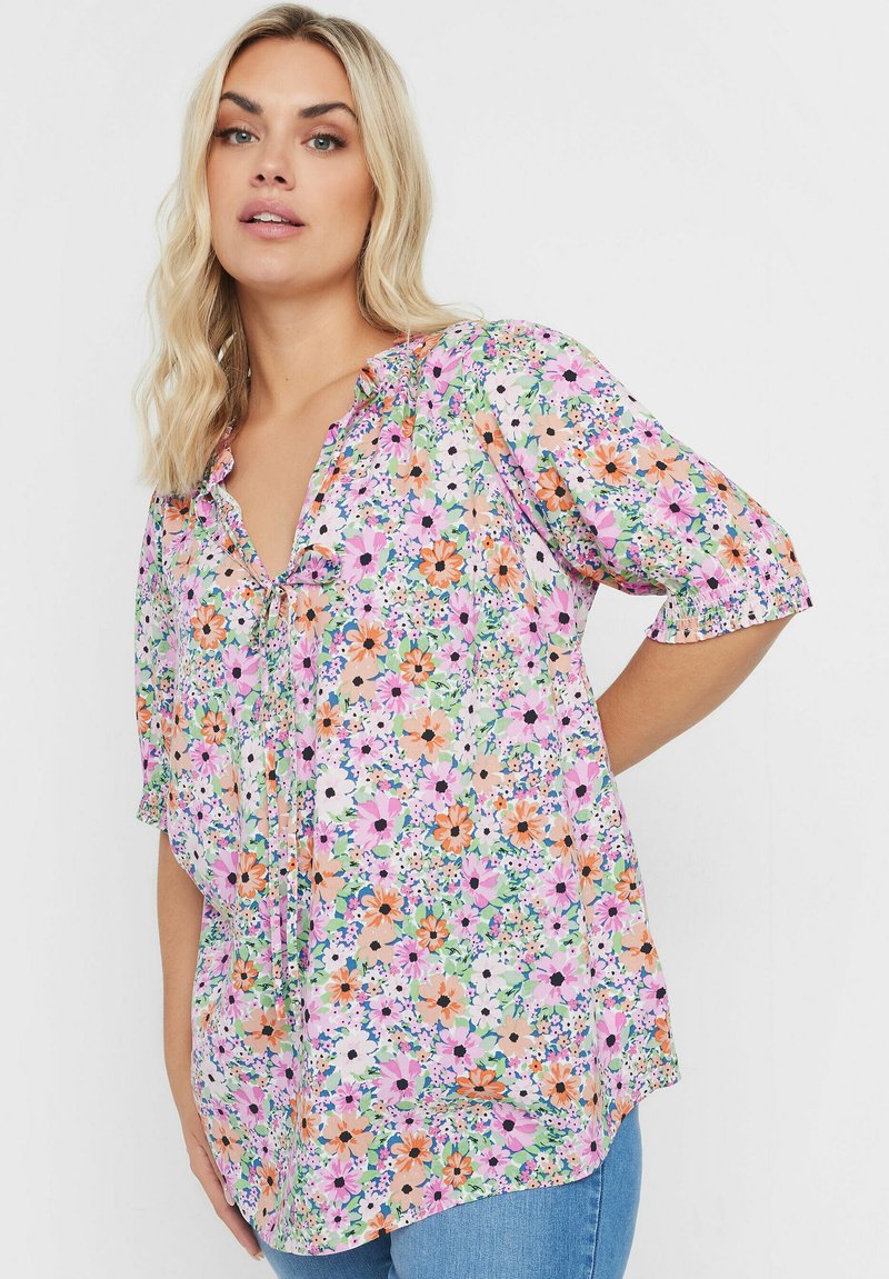 Blommig blus med korta ärmar, med ett flerfärgat blommönster på en ljus bakgrund. Inkluderar en knytning vid halsen och mjuk tyg.