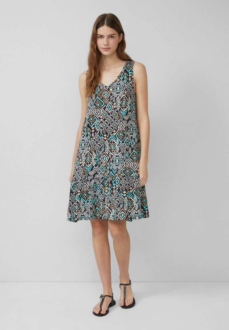 Ärmelloses Kleid in Schwarz mit einem geometrischen Muster in Türkis, Weiß und Braun. Ausgestellter Rock und weicher Stoff. Fußbekleidung: schwarze Sandalen.
