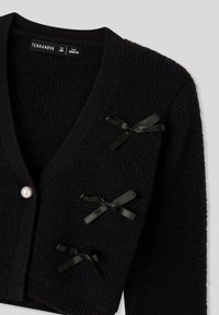 Cardigan nero con design a V, materiale in maglia testurizzata, tre dettagli in nastro nero e un bottone centrale in perla bianca.