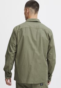 Olijfgroen overhemd met lange mouwen, gemaakt van textured stof, met een traditionele kraag en geknoopte manchet. Etiket zichtbaar aan de rand.