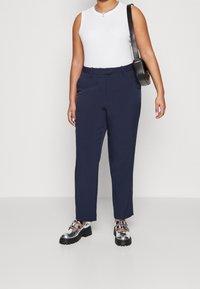 Femme portant un haut sans manches blanc, un pantalon droit bleu marine, des chaussures à boucle en argent, et portant un sac à bandoulière noir sur un fond uni.