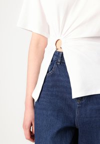 Tricou alb din bumbac, cu un detaliu plisat la spate, având o decupare circulară și accente metalice, purtat cu blugi de denim închis.