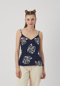 Navyblaue Spaghettiträgerbluse aus leichtem Stoff mit weißem Blumenmuster, V-Ausschnitt und dünnen verstellbaren Trägern, getragen mit hellen Hosen.