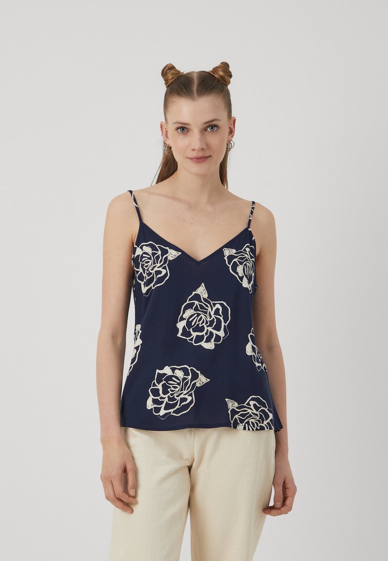 Navyblaue Spaghettiträgerbluse aus leichtem Stoff mit weißem Blumenmuster, V-Ausschnitt und dünnen verstellbaren Trägern, getragen mit hellen Hosen.