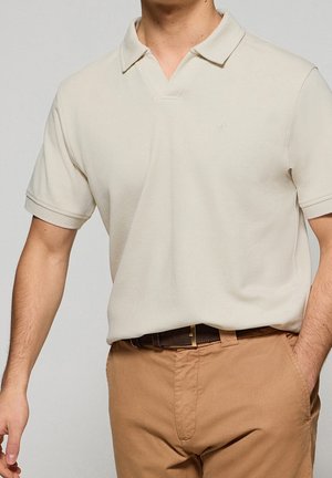 Mężczyzna w jasnobeżowej koszulce polo z krótkim rękawem, włożonej do spodni w kolorze karmelowym, z ciemnobrązowym paskiem, z ręką w kieszeni.