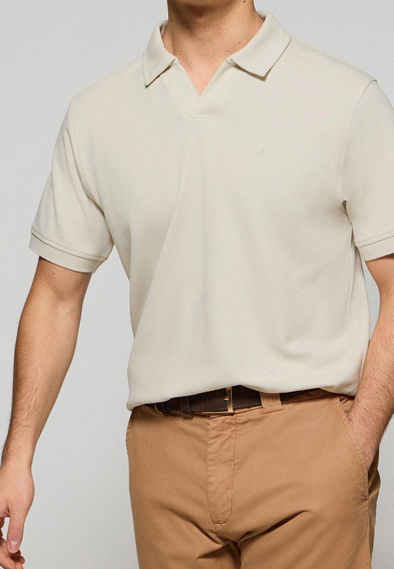 Homme portant un polo à manches courtes beige clair rentré dans un pantalon camel avec une ceinture marron foncé, main dans la poche.