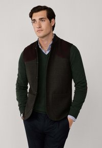 Gilet di lana verde scuro con dettagli in suede marrone sulle spalle, caratterizzato da un motivo a quadri e tasche davanti, indossato sopra un maglione verde a V.