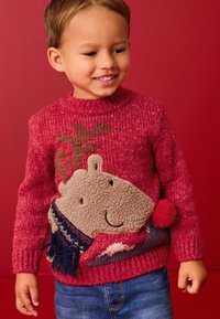 Pull en tricot rouge avec un design texturé d'animal souriant portant une écharpe et un pompon. Assorti à un jean en denim bleu.