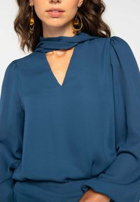Blouse bleue avec un col en V et un détail drapé au niveau du col, dotée de manches longues avec des épaules froncées. Tissu lisse et design élégant.