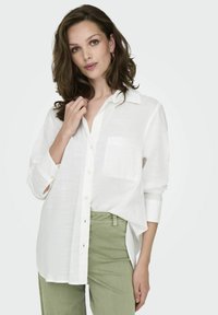 White button-up shirt made of lightweight fabric, featuring a single pocket, rolled sleeves, and a relaxed fit, paired with green pants.