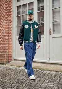 Πράσινο varsity μπουφάν με μαύρα δερμάτινα μανίκια, γκρι φούτερ, μπλε cargo παντελόνι, λευκά αθλητικά και πράσινο καπέλο. Αστική τοποθεσία.