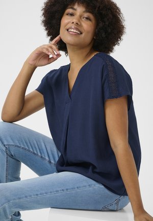 Navyblaues kurzärmeliges Blusenshirt mit V-Ausschnitt, das mit filigranen Spitzenverzierungen an den Schultern versehen ist. Der leichte Stoff fällt weich.