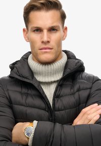 Chaqueta de plumas negra con textura acolchada y capucha, usada sobre un suéter de cuello alto de rayas en gris claro. Reloj dorado en la muñeca con esfera azul, brazos cruzados.