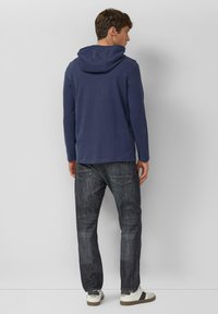 Felpa blu navy con cappuccio e maniche lunghe, abbinata a jeans in denim scuro con vestibilità comoda e due tasche posteriori. Scarpe casual completano l'outfit.