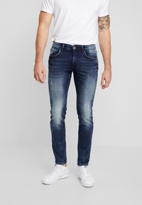 Hombre que lleva unos vaqueros ajustados de un azul oscuro desgastado, zapatillas blancas y una camiseta blanca, de pie frente a un fondo claro y sencillo.