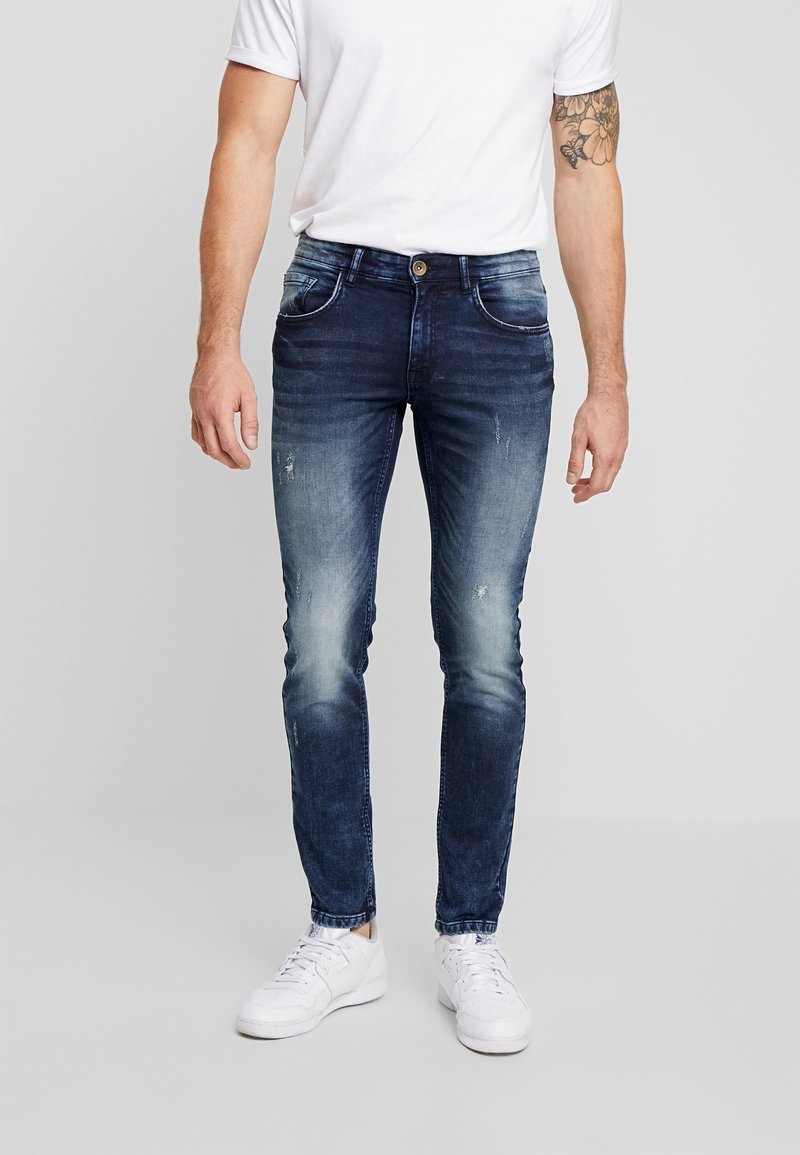 Hombre que lleva unos vaqueros ajustados de un azul oscuro desgastado, zapatillas blancas y una camiseta blanca, de pie frente a un fondo claro y sencillo.