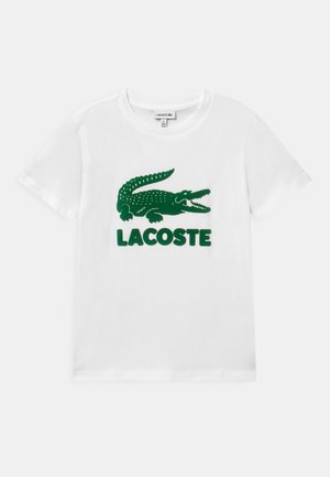 Biała koszulka z krótkim rękawem z dużym zielonym logo krokodyla Lacoste i napisem „LACOSTE” na środku przodu.