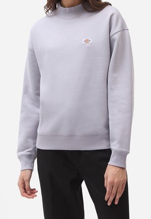 Henkilö, jolla on vaaleanharmaa vetoketjullinen collegepaita, jossa pieni logo rinnassa, ja mustat housut yksiväristä taustaa vasten.