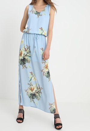Lichtblauwe, bloemen maxi-jurk met een V-hals, elastische taille en zijsplitten, gecombineerd met zwarte sandalen met bandjes.