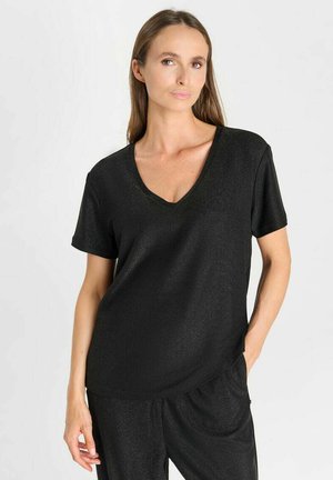 Donna con capelli lunghi e lisci che indossa una maglia nera a maniche corte con scollo a V e pantaloni neri abbinati, in piedi con una mano in tasca.