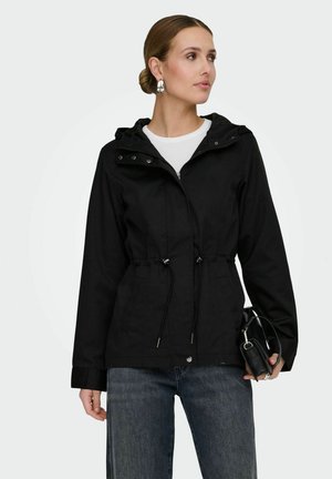 Femme portant une veste noire à capuche avec des cordons, un jean bleu, des boucles d'oreilles argentées en forme de hoop, tenant un sac à main noir, regardant sur le côté.