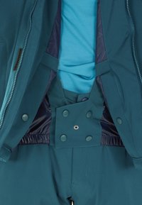 Türkise Jacke und Hose mit innerem, geknöpftem Klappen und darunter liegendem blauen Hemd, die die Details der schichtigen Winteroberbekleidung hervorheben.
